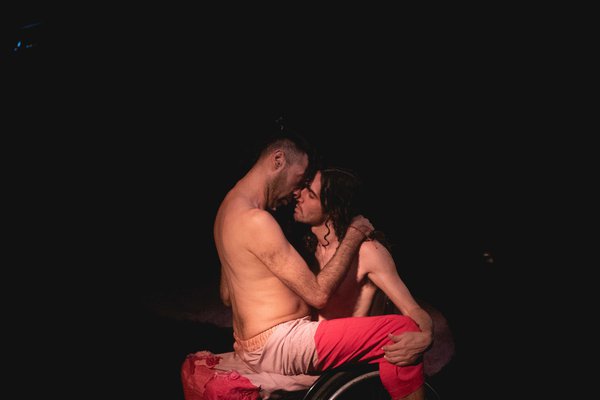 Imagen de fondo negro,  en el centro 2 cuerpos (Javier y Diego), uno de ellos en silla de ruedas, ambos de pantalones rosados, torso y pies desnudos. Están enfrentados uno sentado arriba de otro, abrazados, a un instante de besarse en la boca Imagen de fondo negro,  en el centro 2 cuerpos (Javier y Diego), uno de ellos en silla de ruedas, ambos de pantalones rosados, torso y pies desnudos. Están enfrentados uno sentado arriba de otro, abrazados, a un instante de besarse en la boca